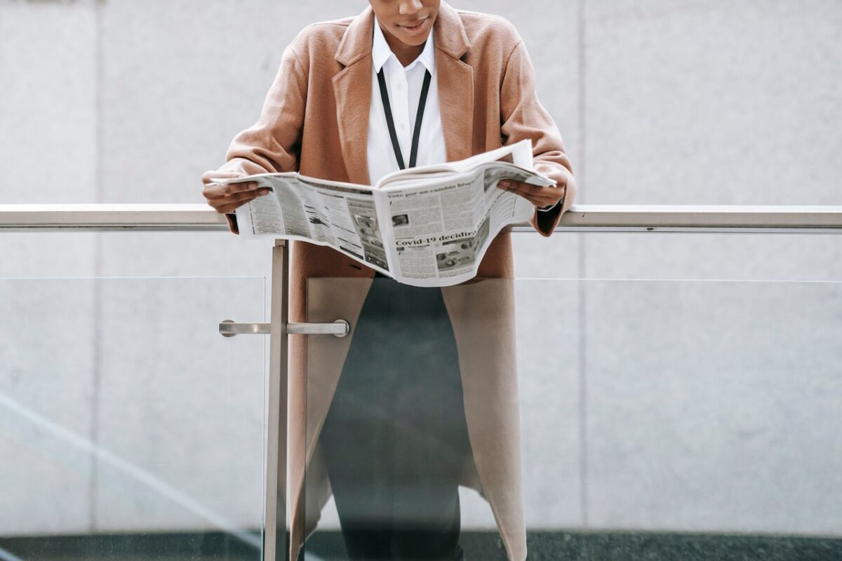 service-01 Crop content African American businesswoman in formal clothes reading fresh newspaper and leaning on glass railing of contemporary office building
