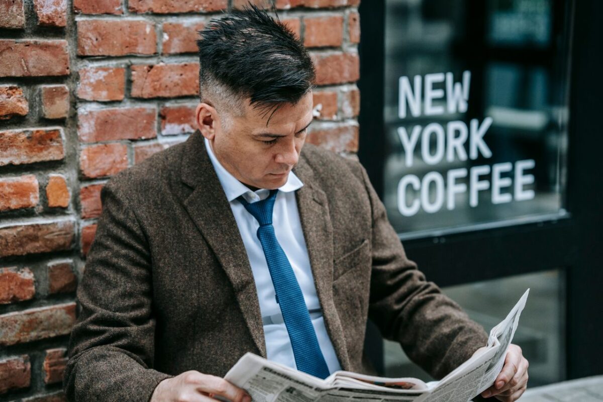 Man in formal attire reading a newspaper outside New York coffee shop.